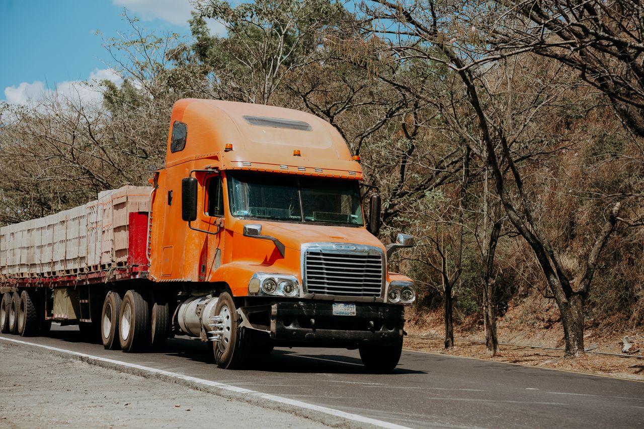 A vibrant orange Freightliner truck drives on a highway through a dry, scenic landscape in El Salvador.